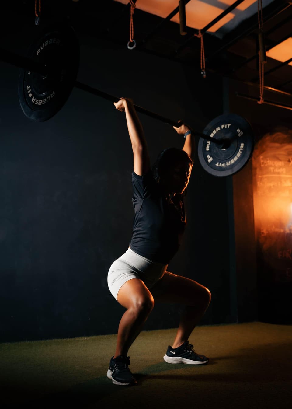 A lifter holding a barbell overhead in the bottom of a squat, lit from the side