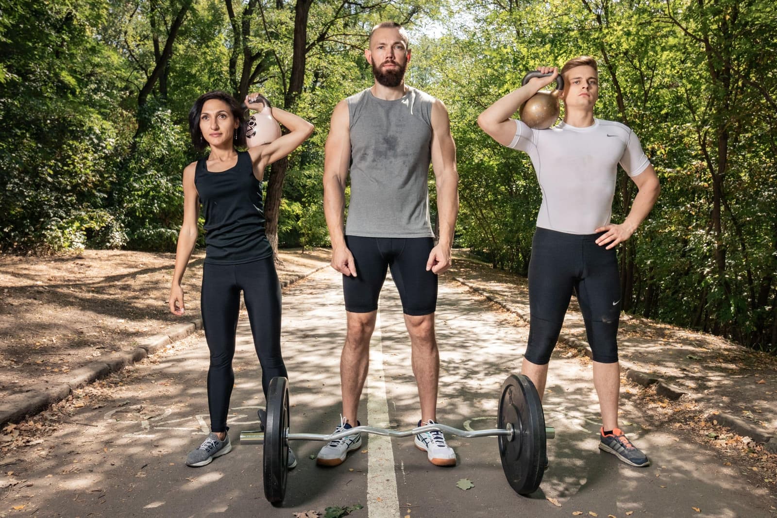 Three people training with a barbell and kettlebells on a park path